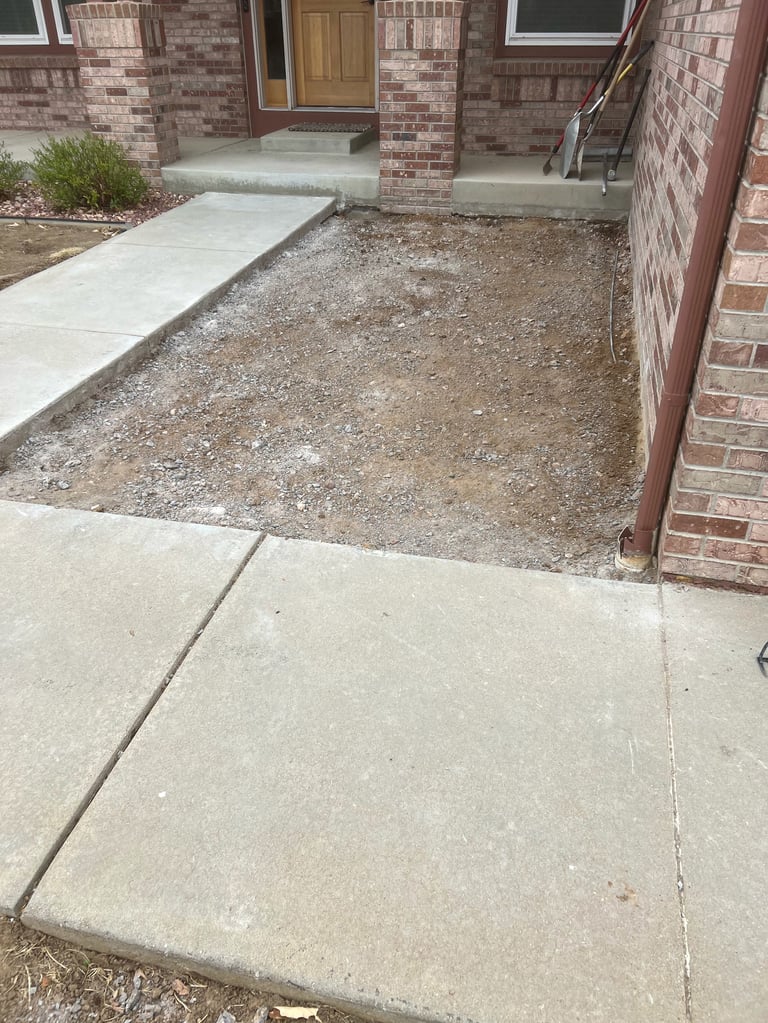 Concrete walkway with dirt and gravel area leading to brick home entrance with door and windows