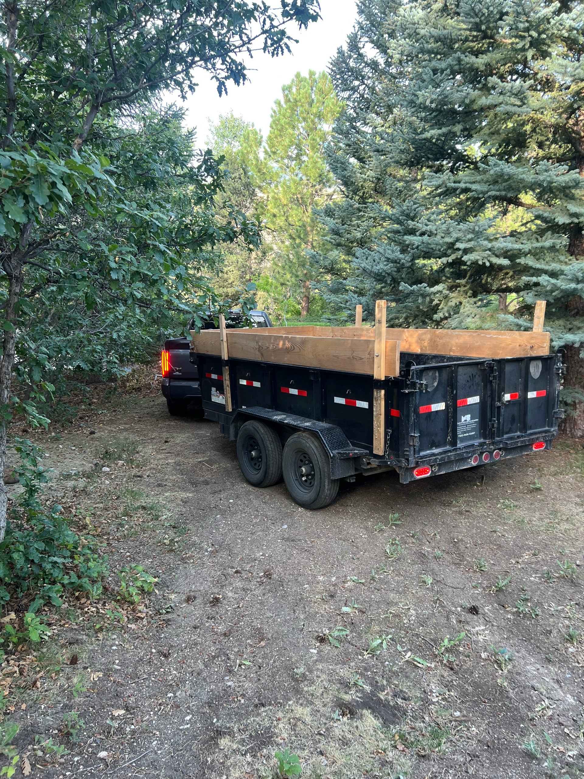 Black dump trailer with wooden sides parked on a dirt road surrounded by trees