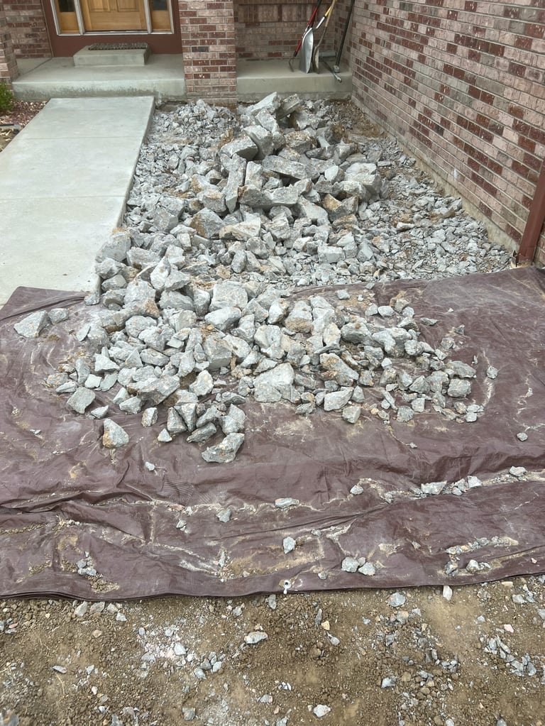 Pile of broken concrete and rubble on dark landscaping fabric beside brick house with driveway